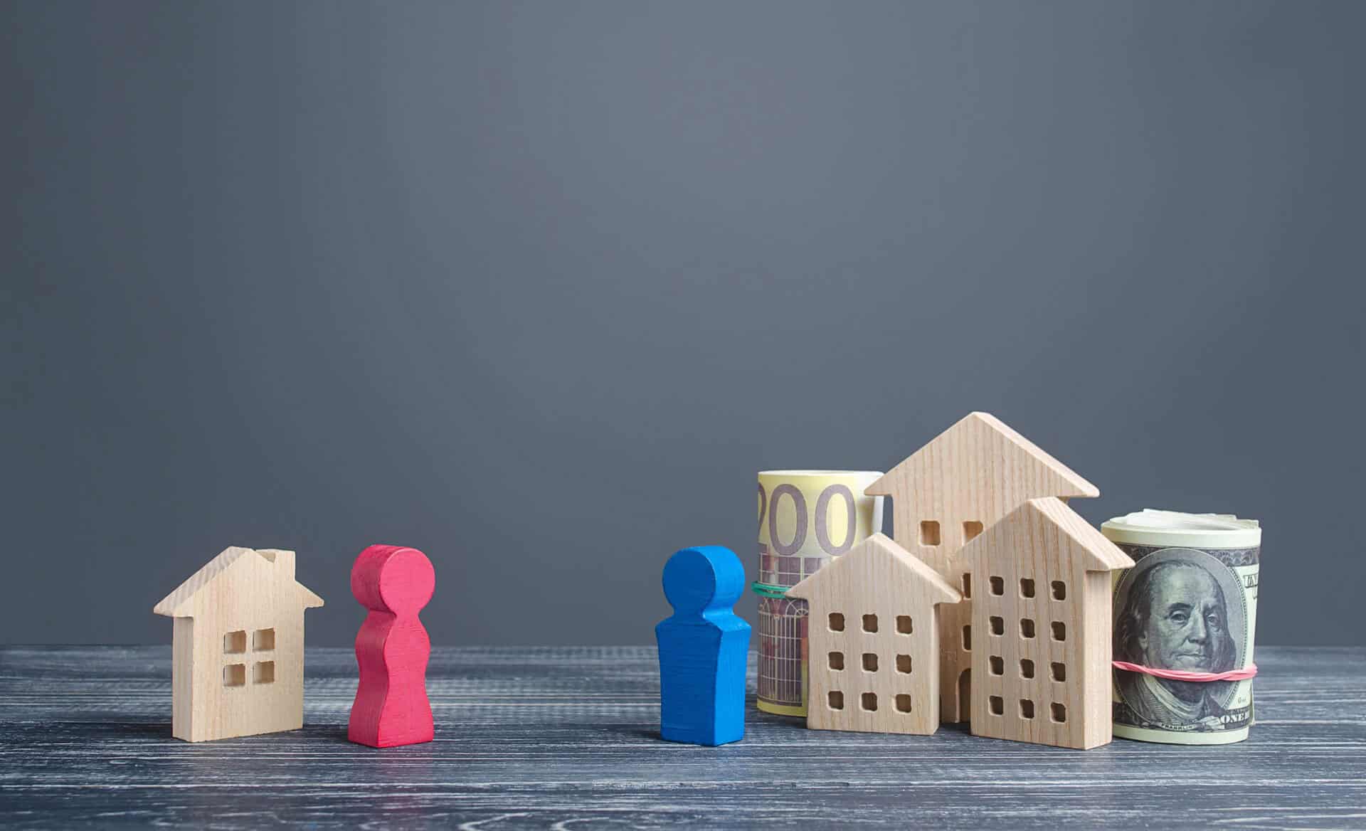 Houten figuren en huizen staan op een tafel, met opgerolde euro- en dollarbiljetten. Twee figuren en een klein huisje staan los van drie figuren, drie huizen en het geld, wat rijkdom en eigendomsverdeling symboliseert.