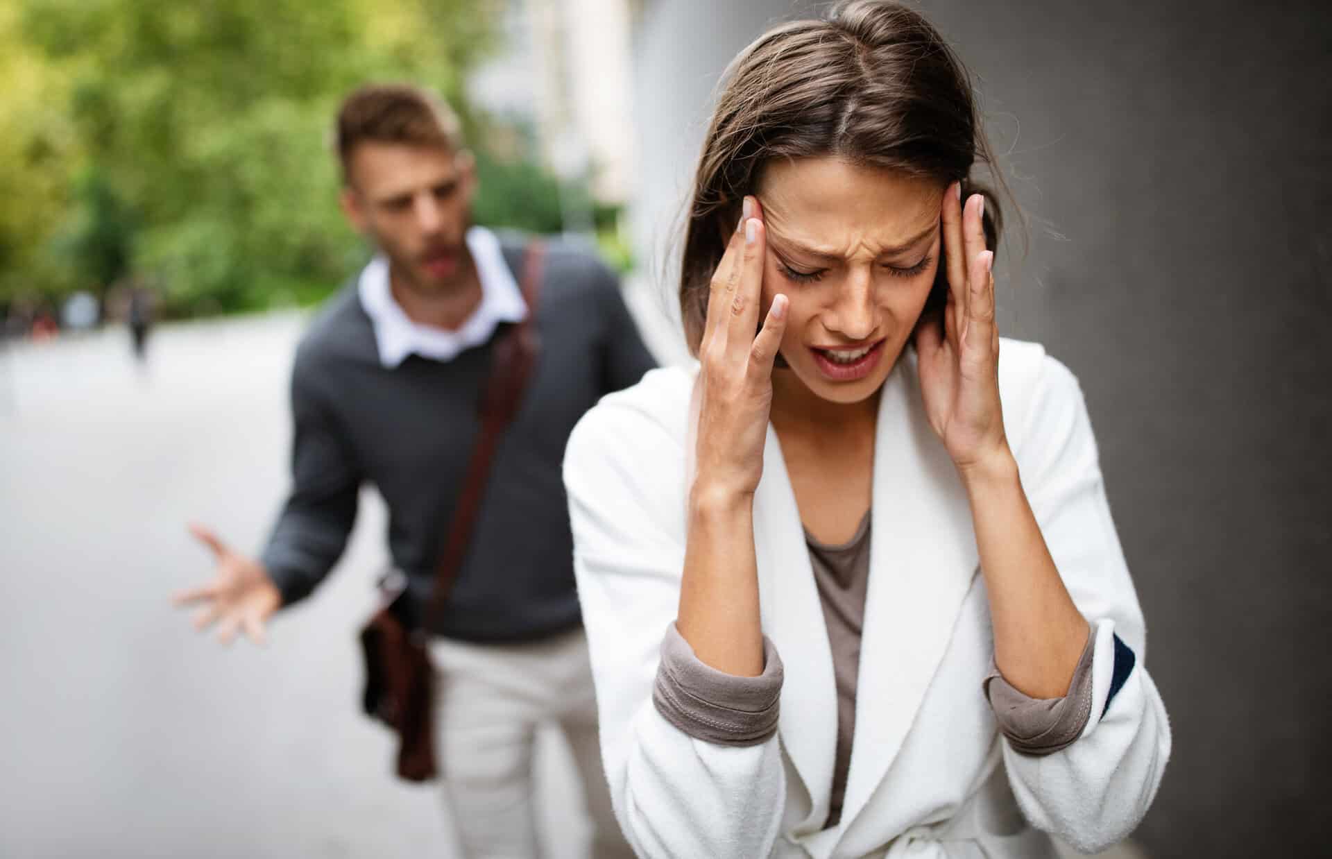 Een vrouw ziet er bedrukt uit en houdt haar hoofd met beide handen vast, terwijl een man op de achtergrond gefrustreerd gebaart en het lijkt alsof hij buiten met haar aan het ruziën is.