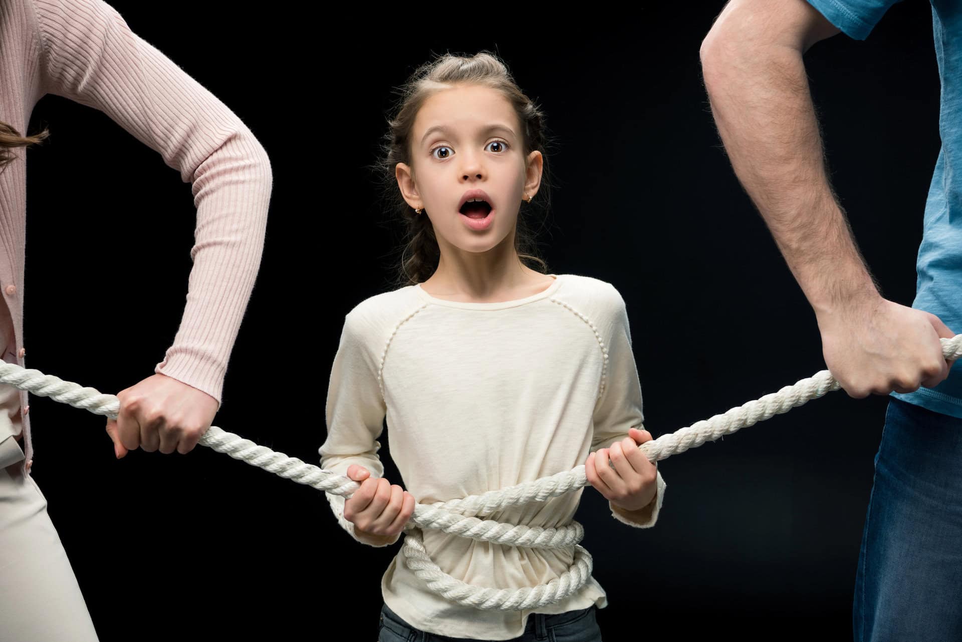 Een verbaasd jong meisje staat met een touw om haar middel gebonden, vastgehouden door twee volwassenen die in tegengestelde richtingen trekken, tegen een zwarte achtergrond.